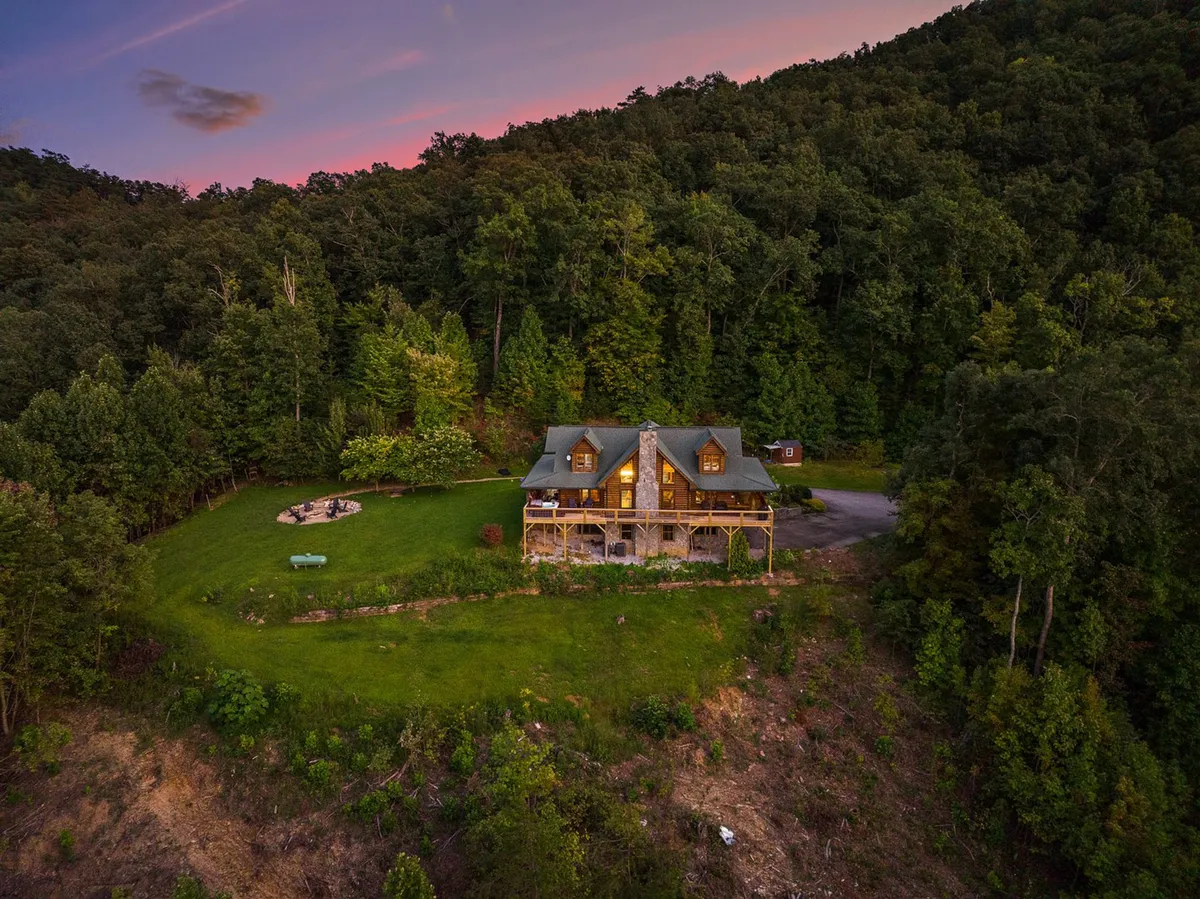 A cozy log cabin nestled in a forested setting with a green lawn, fire pit, and towering trees all around. The open lawn and fire pit area invite gatherings, playtime with pets, or simply soaking in the fresh mountain air. The natural woodland backdrop provides a sense of exploration and proximity to trails.