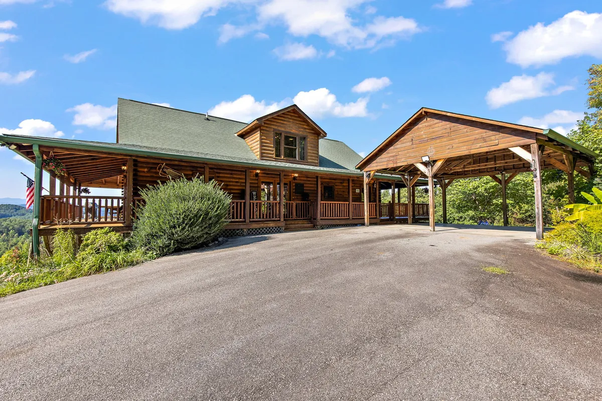 A welcoming log cabin with a wide wraparound porch sits amid green hills, under a bright blue sky dotted with clouds. The expansive porch is perfect for relaxing or letting dogs lounge outside, while the natural surroundings hint at the adventure trails beyond the property.