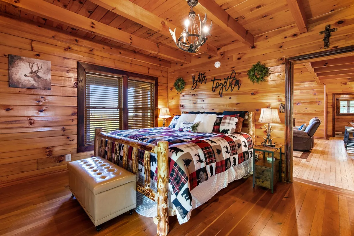 A bright, welcoming bedroom with a wooden bed frame, plaid bedding, warm lighting from a chandelier, and tasteful rustic décor. Nightstands and decorative wall art complete the inviting space, which feels both serene and practical.