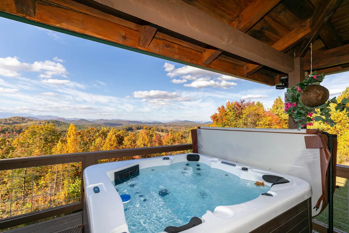 A tranquil exterior shot focusing on the bubbling hot tub set against the mountain view, framed by wooden beams and fall foliage. The atmosphere is one of rejuvenation and celebration, ideal for stargazing or sunset toasts with loved ones.