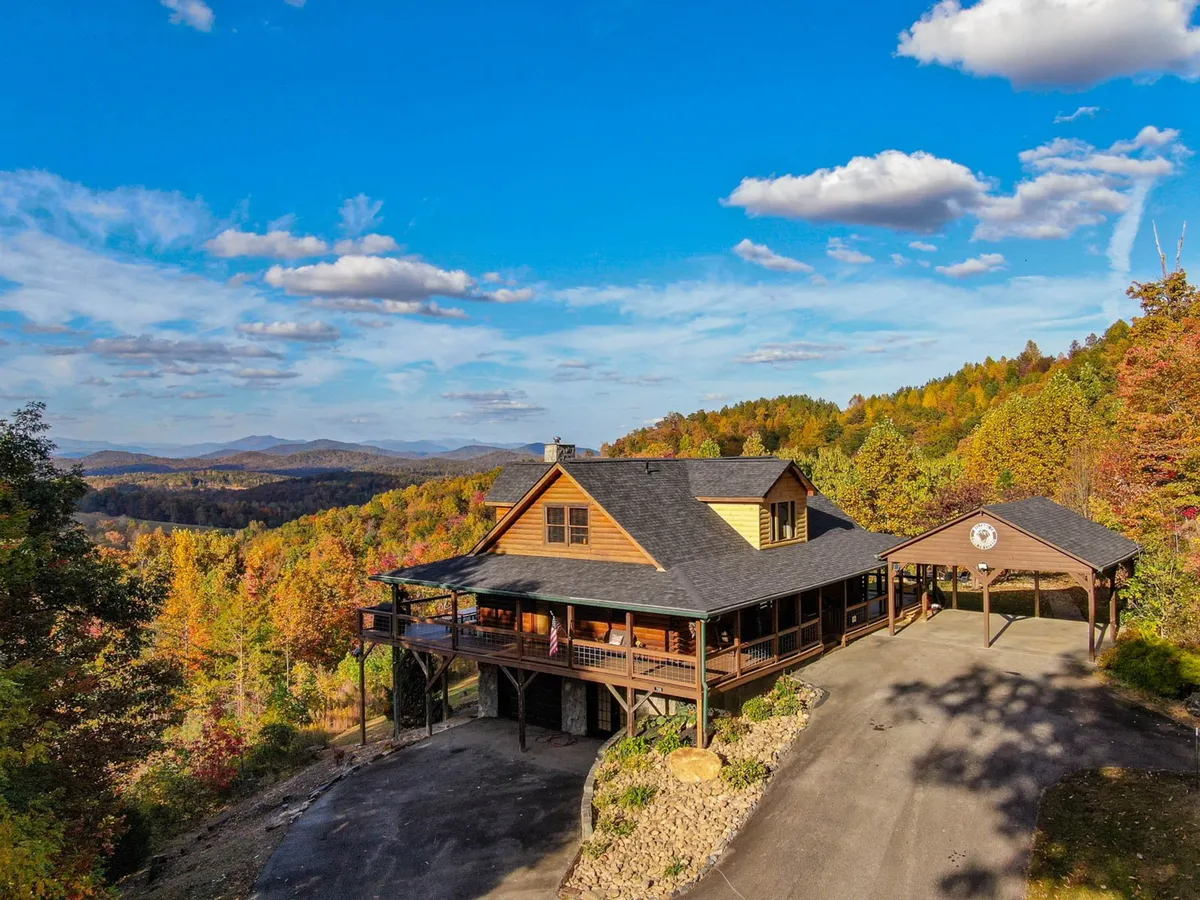 The image showcases the exterior of a spacious log cabin home nestled in the Blue Ridge Mountains. The focus is on the expansive wooden deck, which offers sweeping mountain views, and the inviting architecture of the home. The tranquil surroundings hint at opportunities for outdoor gatherings, peaceful mornings with coffee on the deck, and stargazing at night.