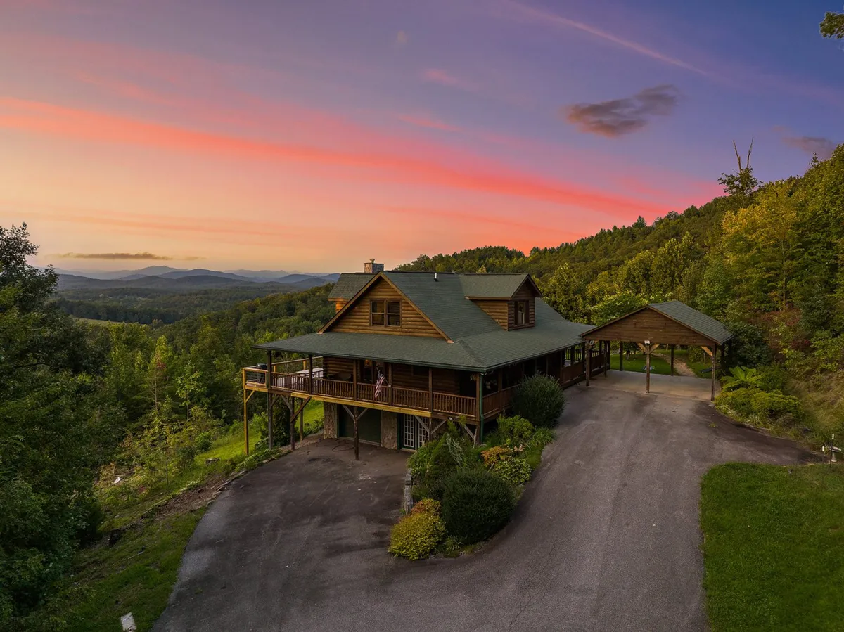 This image captures the log cabin set against a dramatic backdrop of surrounding mountains and a colorful sunset sky. The expansive deck with railing is visible, inviting guests to relax outdoors and soak in the panoramic views. The scene evokes tranquility and the sense of being immersed in nature.