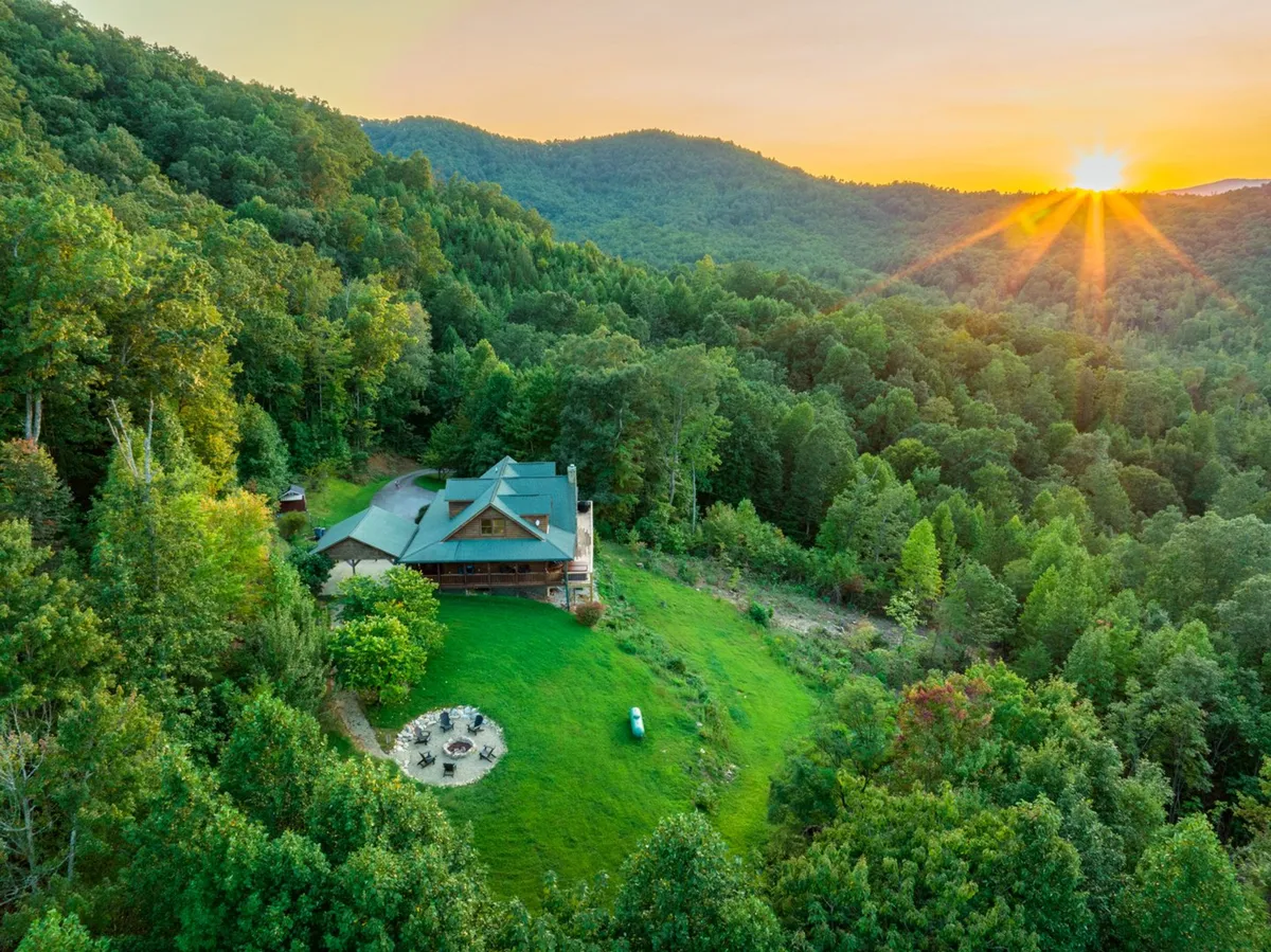 The exterior of a charming log cabin surrounded by trees and mountains, with a welcoming fire pit setup in the yard. The scene suggests space for outdoor gatherings, campfires, and enjoying nature together as a family.
