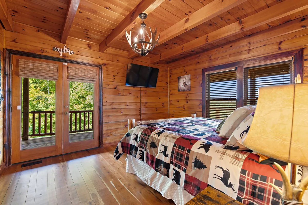 A rustic master suite featuring a plaid bedspread on a wooden bed frame, antler chandelier overhead, and double doors opening to a private balcony. The room is cozy, spacious, and well-lit, with warm wood tones and a television mounted on the wall.