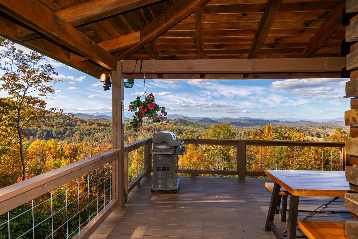 An inviting exterior shot of the wrap-around deck, complete with a grilling area, dining table, hanging plants, and breathtaking mountain views. The scene is set for outdoor dining, relaxing, and enjoying the panoramic vistas that define the home’s unique setting.
