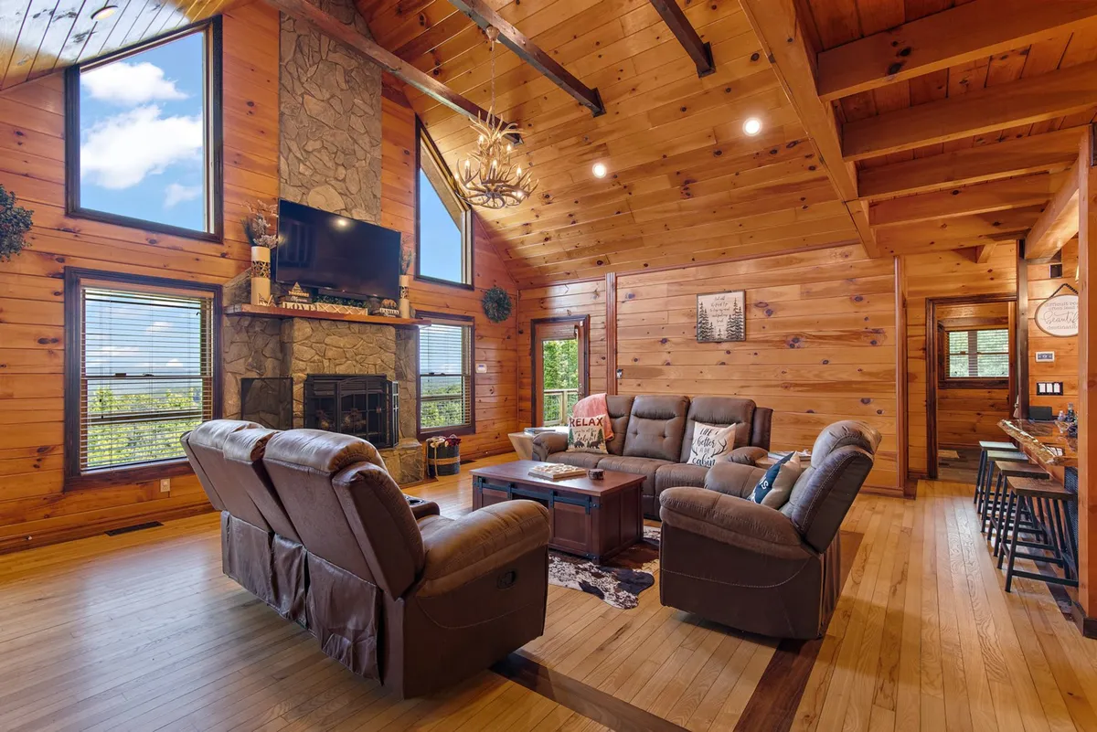 This image captures the spacious open-concept living room, featuring a striking stone fireplace, large windows framing the Blue Ridge Mountain vistas, exposed wooden beams, and inviting comfortable seating. The natural light and warm decor create a welcoming atmosphere for group gatherings and relaxation.