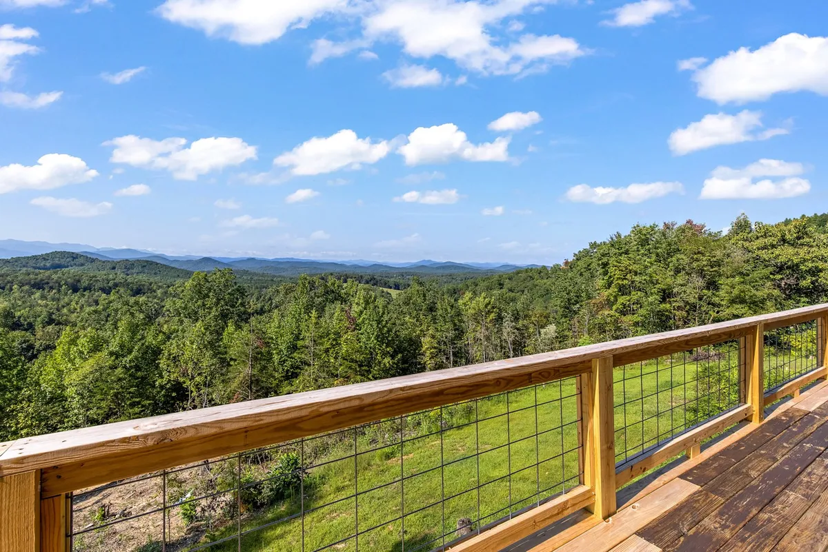 A breathtaking mountain range stretches across the horizon, viewed from behind a wooden deck railing. Lush green forests extend below, and the peaceful mood captures the essence of relaxation and immersion in nature. The scene is ideal for wildlife watching, photography, or simply unwinding in awe of the landscape.