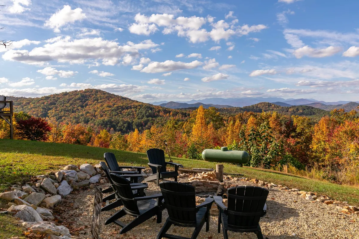 This outdoor scene features a stone fire pit encircled by Adirondack chairs and overlooks mountain views decked with fall foliage. The setting is ideal for evening gatherings, stargazing, and fireside chats under the sky.