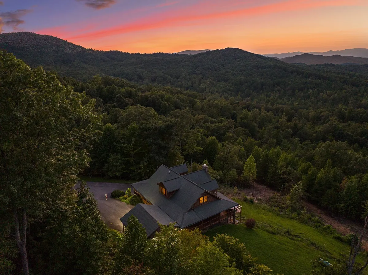 A serene wide-angle shot showing the house nestled amidst a forest and set against a mountain backdrop at sunset. The space looks ready for hosting memorable events, outdoor games, or simply admiring sunset views with loved ones.