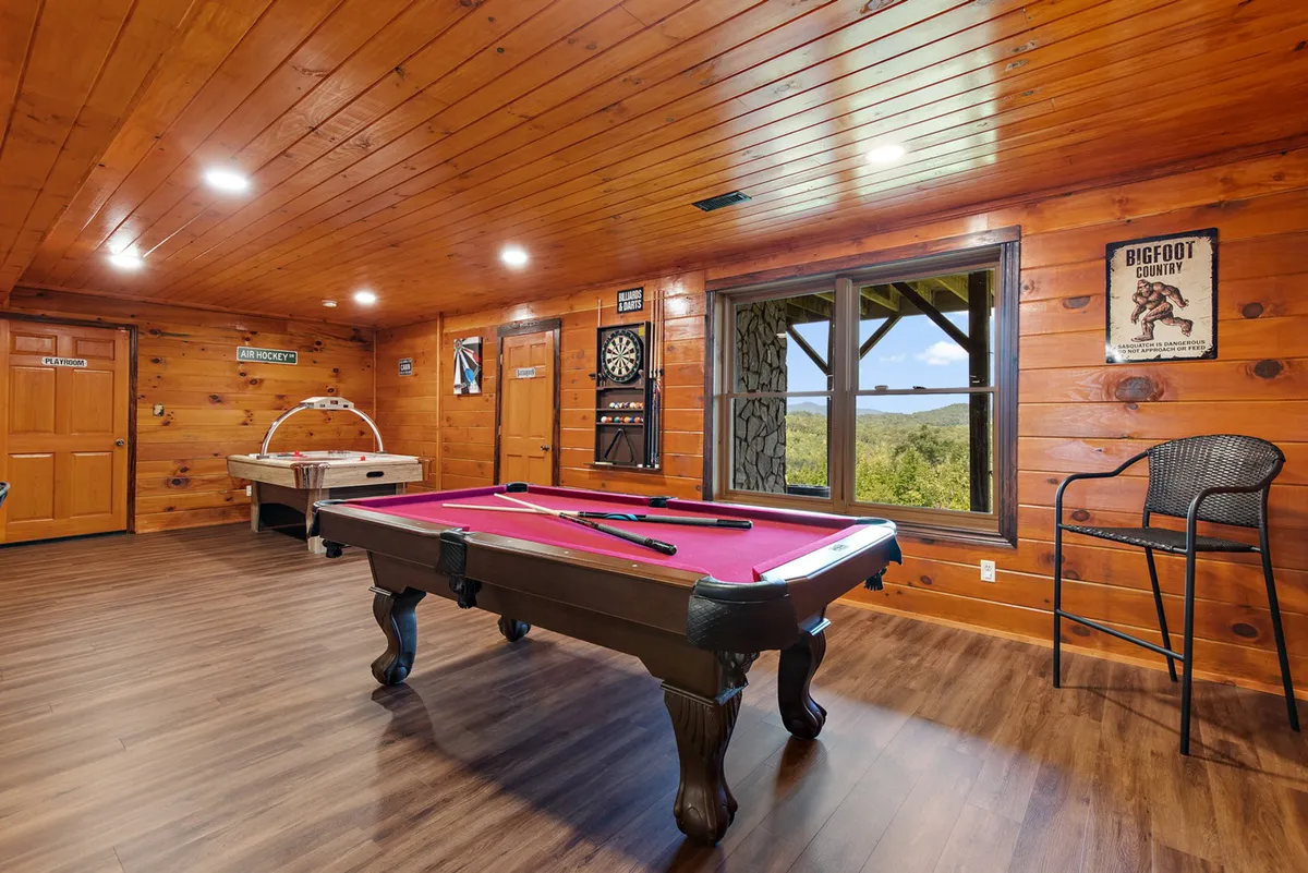 This image features a lively game room with a pool table at the center, a dart board on the wall, and an air hockey table by a large window that offers pleasant views. The space is designed for recreation, with ample room for players and spectators alike, and bathed in natural light for a bright, energetic atmosphere.