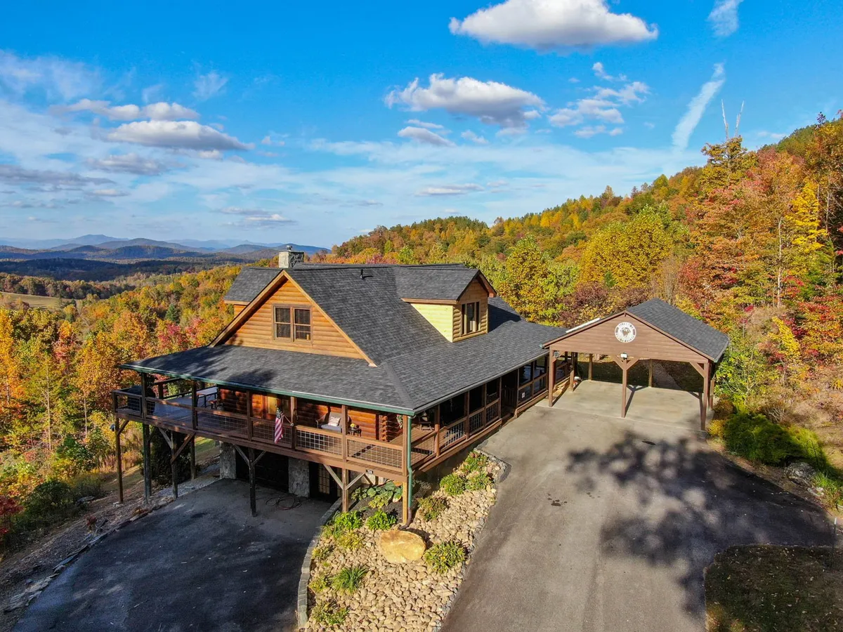This image shows the log cabin-style home with an expansive porch, surrounded by autumn foliage and featuring the carport. The scene is relaxed and inviting, with visible ample parking and the covered carport, both important amenities for adventurous guests and their families—including those with pets.
